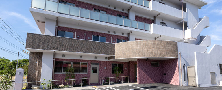 介護施設の建物外観。白と赤褐色のタイル張り、複数階建て、駐車スペース付き